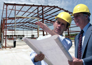 Two men in hard hats with plans stand before a prefab metal building frame.