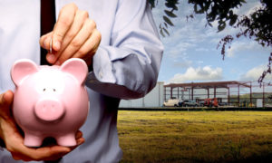 Photo of a man putting money in a piggy bank as he stands before his metal building under construction.