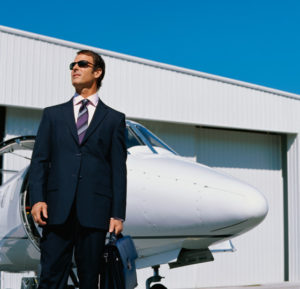 Photo of a man in a suit standing by a corporate jet in front of a steel airplane hangar.