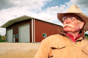 Rancher stands before a RHINO red metal barn with white trim.